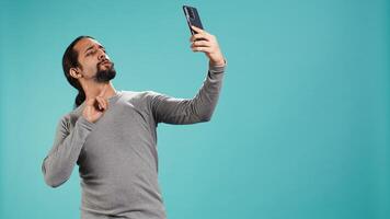 Man using smartphone to take selfies and post them on social media, studio backdrop. Vain person taking photos using phone selfie camera, isolated over studio background, camera B