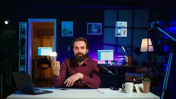 Tech guru in dimly lit apartment studio filming smart light bulb review for online streaming platforms. Media star hosting show, presenting IoT home automation product, camera B photo
