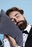 Hotel staff takes quick nap using pillow in studio, feeling extremely tired and worn out. Young man working as bellhop falling asleep standing, indicating fatigue and burnout. photo
