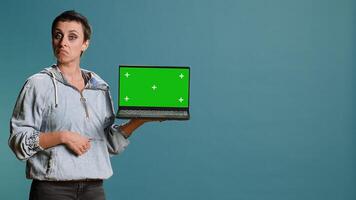 Young adult pointing at laptop with isolated chroma key screen in studio, using modern technology on wireless gadget. Woman with confident style does an advertisement. Camera B. photo