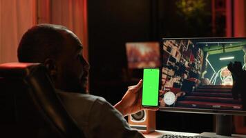 Isolated screen smartphone held by player competing in online multiplayer videogame at home. Mockup mobile phone utilized by person playing on gaming PC in neon illuminated apartment photo