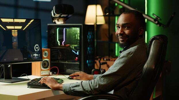 Portrait of smiling man playing FPS videogame on PC screen in neon illuminated apartment living room. Cheerful gamer enjoying shooter game on computer desktop display in RGB lit home, camera A photo