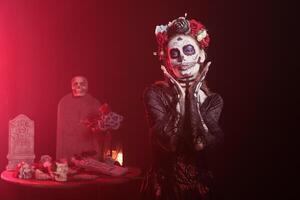 Woman in occult decorated studio during Day of the Dead Halloween event honoring dead loved ones. Scary lady doing ancient Aztec custom of celebrating ancestors, red light background photo