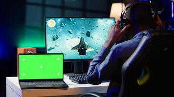 Mockup laptop next to man playing high FPS singleplayer videogame with spaceship shooting lasers. Isolated screen notebook near gamer using crosshair to hit targets in SF game photo