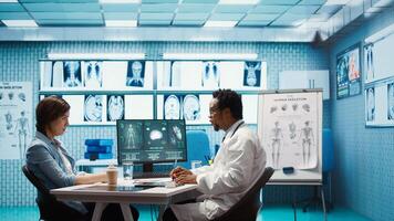 Male physician doing an x ray diagnostics scan interpretation during check up visit, discussing about recovery treatment in cabinet. Doctor examining radiography test results for a patient. Camera B. photo