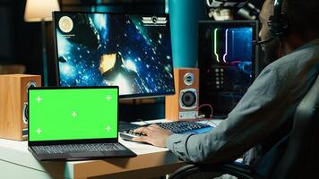 Isolated screen laptop next to professional streamer competing in online multiplayer videogame, streaming live at home. Mockup notebook and man playing on PC, using headphones to talk with audience photo