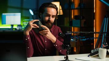 Tech guru presenting V mount battery, filming technology review using cellphone on tripod and professional microphone. Caucasian vlogger showcases charging device specs, recording with phone, camera A photo