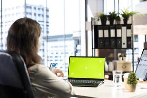 Business director working in her own office with green screen, looking at device display and developing a new project idea. Female entrepreneur empowered by corporate culture. photo