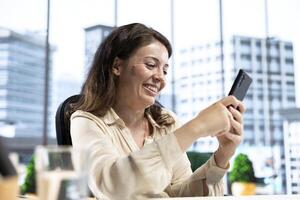 Pleased empowered businesswoman texting messages in her own director office, having fun on social media. Leader discussing with partners about new project ideas for career growth. photo