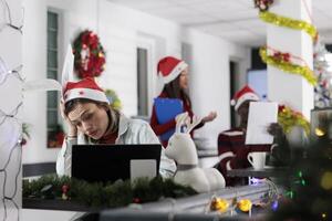 Office employee wearing Christmas hat feeling burnout while struggling to keep up with incoming company project deadline. Worker frustrated by paperwork overload during festive holiday season photo
