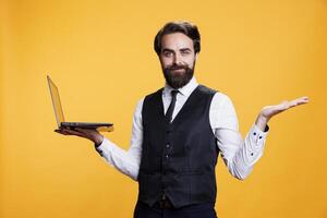 Cheerful employee working on laptop against yellow background, using online connection to develop restaurant website for orders. Professional waiter holds modern pc on camera. photo