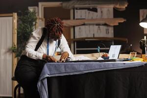Expert focused suitmaker creating bespoken sartorial fashion collection surrounded by sewing materials and sketch drawings. African american couturier working in exquisite tailoring studio. photo