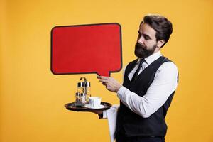 Professional butler holds speech bubble on camera, presenting isolated red empty billboard to create advertisement in studio. Young man waiter shows blank copyspace cardboard sign for ad. photo