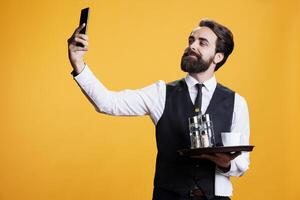 Smiling person taking pictures with phone, working in restaurant industry with tray of cutlery in studio. Professional butler takes photos and selfies on break, using smartphone for fun.
