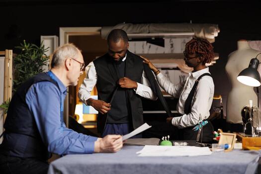 Stylish gentleman trying on custom made suit before needed adjustments are being made by professional suitmakers. Expert tailors working on comissioned bespoke attire for client photo