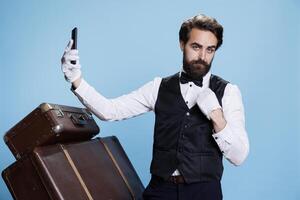 Luxurious hotel worker takes pictures while he is posing in front of camera, feeling confident wearing suit and gloves. Professional elegant doorkeeper using mobile phone app for photos. photo