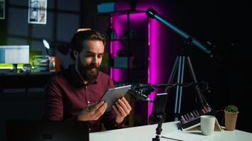 Tech specialist presenting tablet in entry level low budget price range to interested viewers, filming with smartphone on tripod. Man reviewing digital device, recording with mobile phone, camera B photo
