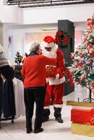 Visitors enter holiday prize draws, worker disguised as Santa Claus inserts cards with names written inside box. Older people waiting in festive store for contest results to win free attire. photo