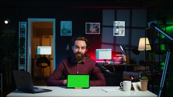 Smiling man in studio filming chroma key tablet review for tech enthusiasts. Cheerful online star hosts technology internet show, unboxing green screen digital portable device, camera B photo
