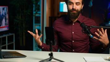 Influencer talks in front on smartphone attached to tripod and high quality mic in studio, discussing with viewers. Man using mobile phone and professional gear to record social media , camera A photo