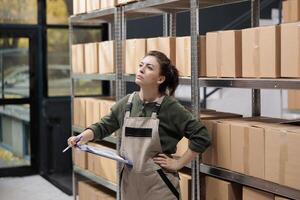 Delivery service employee searching clients parcel in warehouse checking clipboard with online order. Storage room worker wearing uniform overalls supervising goods in factory storehouse photo