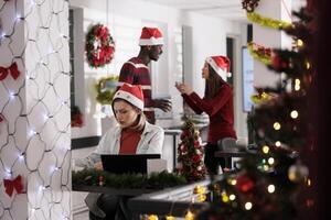 Stressed employee struggling with overdue task deadlines in Christmas decorated workspace while coworkers have fun in background. Businesswoman working in festive themed office during holiday season photo