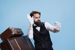 Silly doorkeeper acting playful in studio, feeling cheerful and confident with groovy dance moves in suit. Man working as bellboy being funny and playing around over blue background. photo