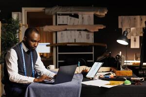 African american suitmaker looking online for outfit inspiration, comparing results with sketch drawings of sartorial garments. Senior couturier ordering sewing materials on laptop in tailoring studio photo