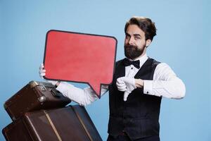 Stylish doorman holds red speech bubble and giving thumbs down in blue studio, creating marketing ad on empty cardboard sign. Hotel porter using empty isolated board, feeling displeased. photo
