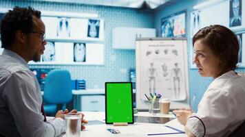 General practitioner pointing at green screen mockup on device, discussing with her patient about recovery treatment. Doctor doing checkup visit on insurance with a man. Camera B. photo
