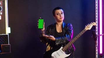 Woman rock musician playing guitar next to green screen display on device, doing a live music performance on stage at the nightclub. Artist rocker acting cool and rebellious with edgy style. Camera A. photo