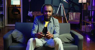 Player in RGB lit home putting headphones on to be immersed in videogame experience, enjoying relaxation time. Man wearing headset sitting on couch, playing games on console using gamepad, camera A photo