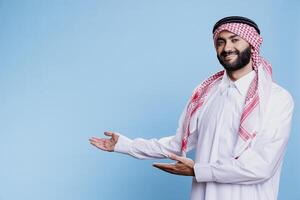 Arab man in thobe and ghutra pointing to side with hands, advertising product studio portrait. Muslim person wearing traditional clothes showing to right direction with arms photo