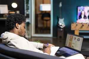 Man working from home doing math calculations using statistical data financial graphs on laptop. Remote employee in living room comparing business charts economic figures, writing on notepad photo