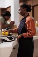 Female farmer vendor in zero waste shop at checkout counter processing customer order. Young client buying organic nutritious food from her ecofriendly local neighborhood store. photo