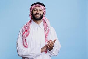 Smiling muslim man showing applause gesture and looking at camera with excited expression. Cheerful arab wearing islamic thobe and ghutra applauding at performance studio portrait photo