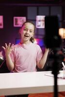 Joyful gen Z internet star doing livestream using cellphone on professional recording tripod. Happy cute kid does live broadcast using phone camera to talk with online audience photo