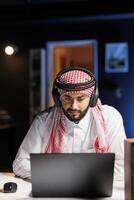 Arab businessman in traditional attire diligently works in office, using laptop for research, browsing, and communication. Muslim guy uses earphones to enable calls, utilizing modern technology. photo