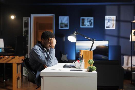 Sleepy african american person dealing with burnout while he works from home, trying to solve business tasks with global internet statistics. Self employed guy being tired at his workstation. photo