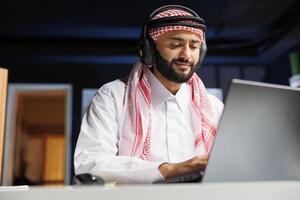 Close-up shot of dedicated Arabian businessman in traditional attire working diligently at his modern desk, utilizing wireless technology and online communication for research and conferencing. photo