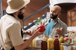 Middle eastern customer inquires about red bell pepper that a male storekeeper with a hat is holding. Vendor and client interacting over group of various glass jars and bottles in grocery store photo