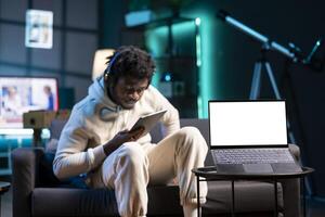 Man reading eBook from tablet in neon lit apartment, focus on mockup laptop. Relaxed person wearing headphones, enjoying lecture sitting on couch next to isolated screen device on coffee table photo