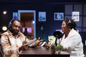 African American podcast host interviews a female content creator in a neon-lit home studio using advanced recording equipment. Their engaging discussion is broadcast digitally to their online fans. photo