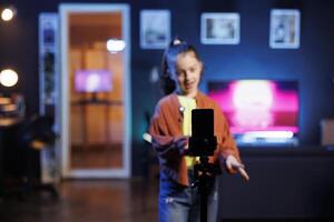 Focus shot on cellphone on tripod used by girl dancing and singing, shooting for social media. Kid in blurry background entertains subscribers with professional choreography recorded with phone photo