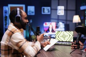 Male talk show host holding a notebook and interviewing a guest, having online discussion for their listeners. African american man using recording equipment to create a radio program at home. photo