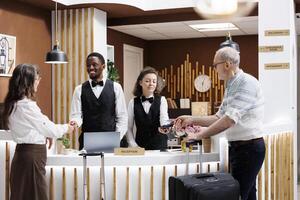 Elderly couple at hotel reception where friendly staff assist with check-in and credit card payment. Comfortable accommodation, convenient procedure at registration counter with retired senior adults. photo