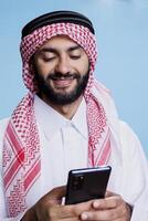 Smiling arab man wearing islamic traditional clothes typing message on smartphone. Muslim person dressed in thobe and ghutra chatting in social media using mobile phone with cheerful expression photo