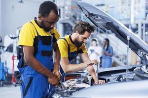 Certified servicemen in auto repair shop working together on fixing car, discussing best options. BIPOC worker and colleague collaborating on servicing broken vehicle, checking for faulty components photo