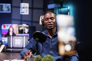 Male guest speaking into microphone, answering questions while looking at phone camera. African american man using mobile device on tripod to record an online talk show at home. photo