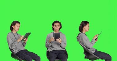 Asian man navigating internet on tablet in greenscreen studio, using mobile device to search through webpage online. Person sitting on chair and working on gadget, relaxed guy. photo